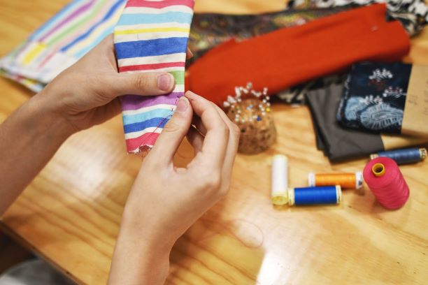 a pair of caucausian hands holding colourful striped fabric and sewing. In the background are scattered fabrics and threads.